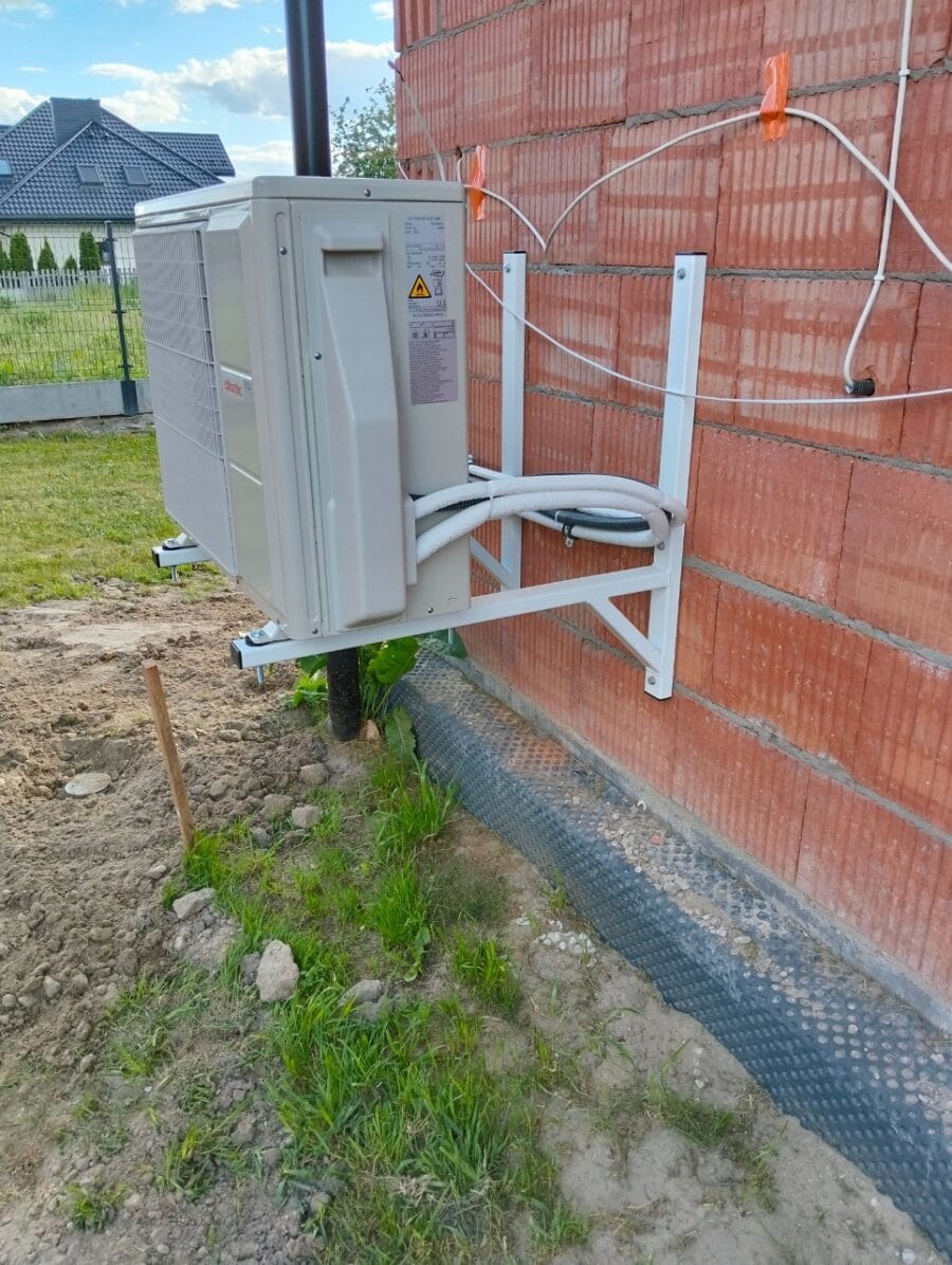 Klimatyzator-ścienny-metal-Serwis-Obrzycko Zewnętrzny klimatyzator ścienny zamontowany na metalowej konstrukcji przy ścianie budynku. Widoczne przewody i elementy montażowe - ogrodzenie Met.